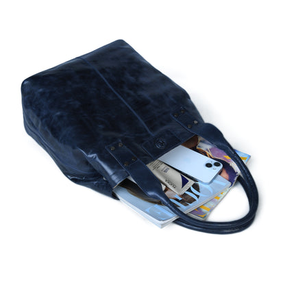 A dark navy leather tote bag shown open on a white background, filled with magazines, a phone, and other items.